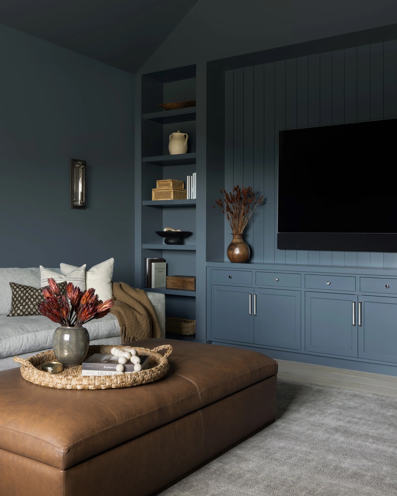 A moody media room with blue-gray built-ins, a tan leather ottoman, and warm accents.