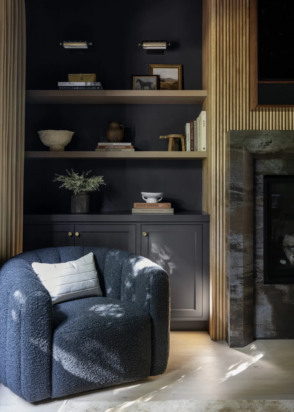 A dark, paneled reading nook with floating wood shelves, a boucle armchair, and brass cabinet hardware.