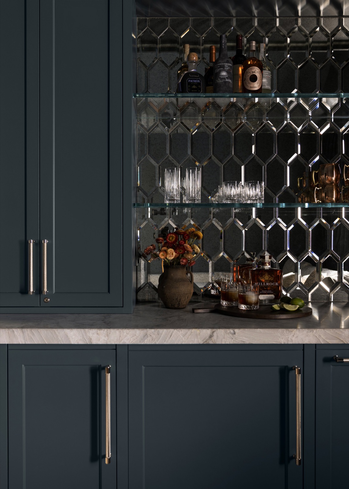 A bar detail, marble counter with dried flowers and a whiskey tray against antiqued mirror tile.