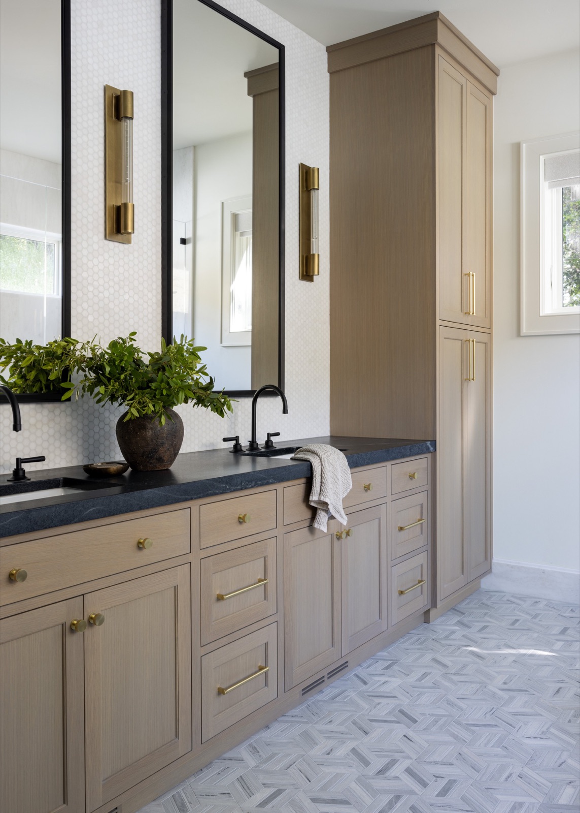 Bath vanity in warm oak with brass sconces and a dark stone counter.